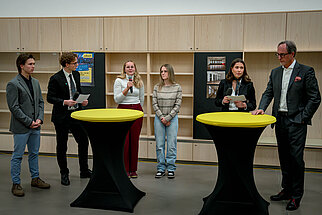 Diskussionsteilnehmerinnen und -teilnehmer bei Veranstaltung in Österreich mit Stehtischen und Regalen im Hintergrund.