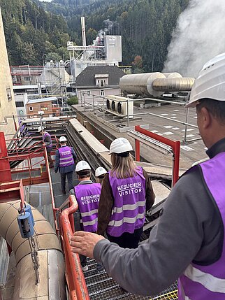 Besucher mit Warnwesten und Helmen besichtigen ein Industrieareal mit rauchenden Schloten.